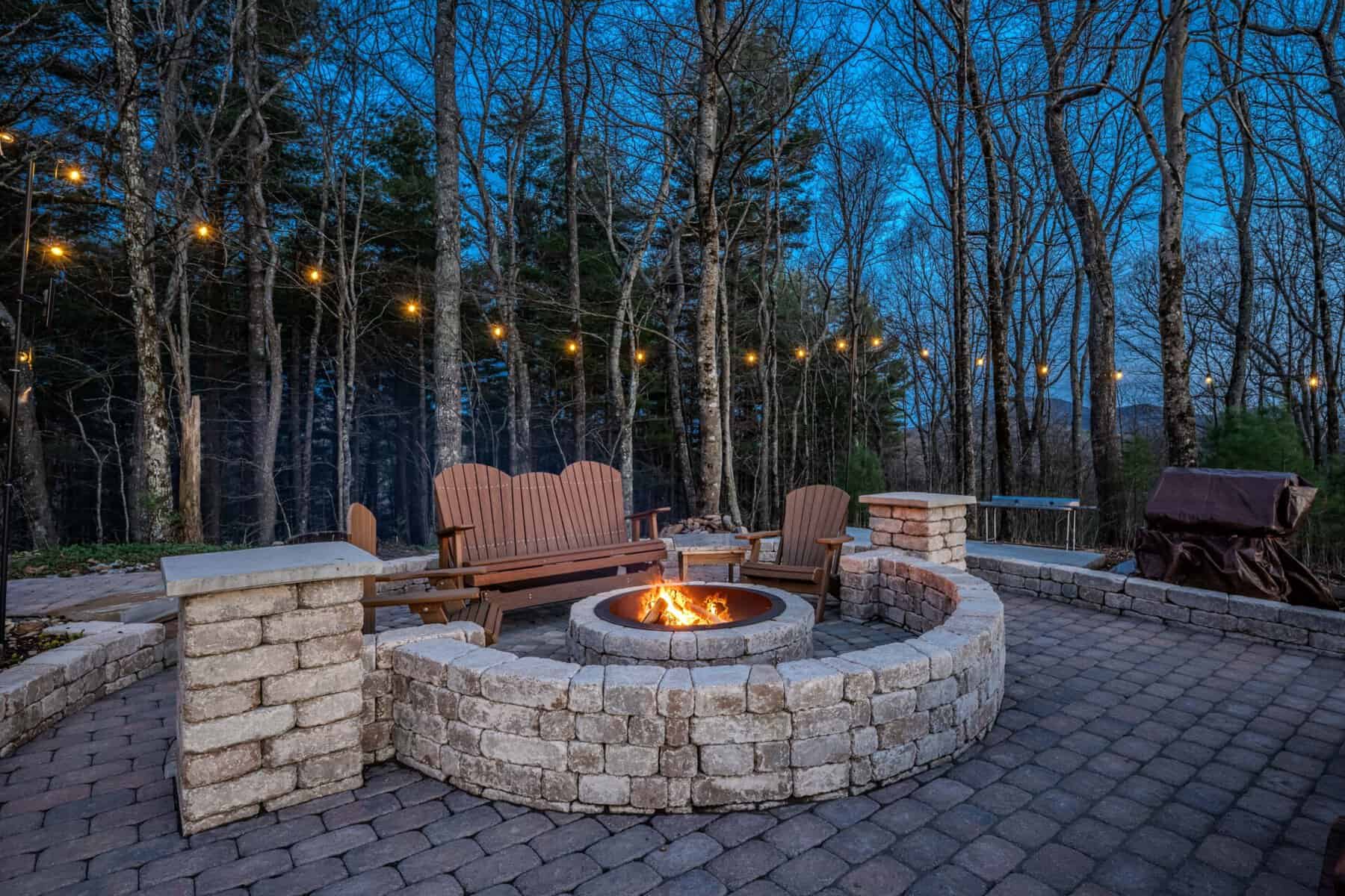 Stone fire pit with wooden benches and string lights in backyard.